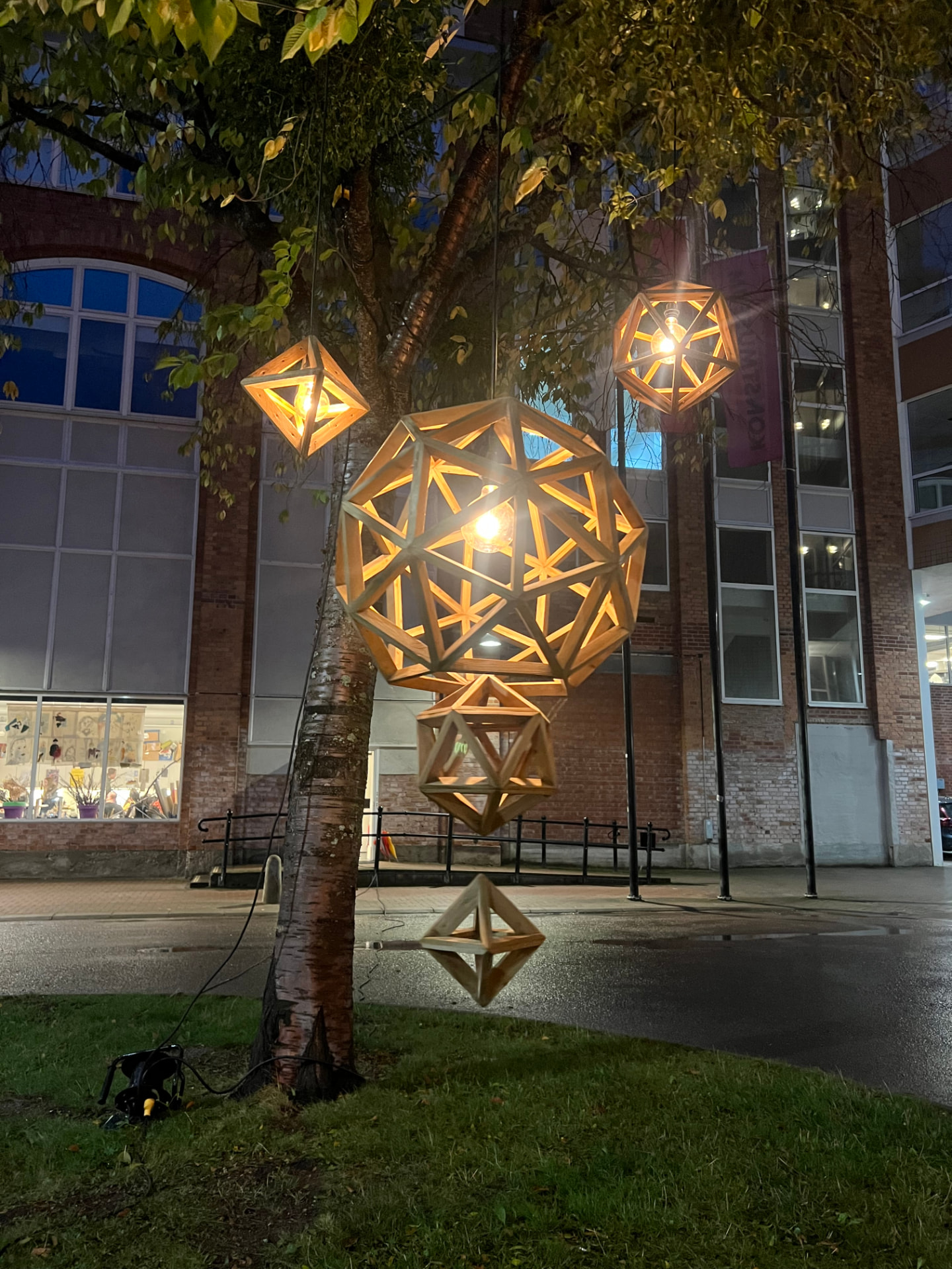 Geometriska träinstallationer upplysta i ett träd utanför en tegelbyggnad kvällstid
