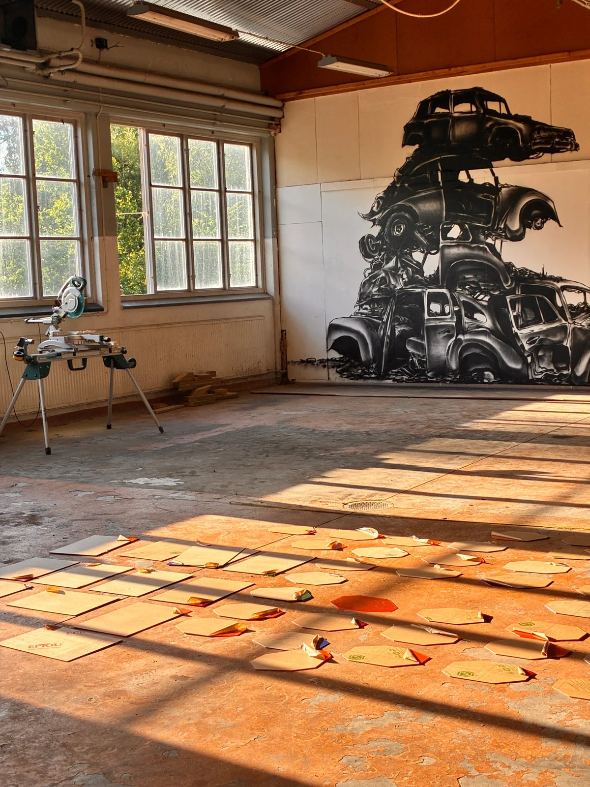 Stor svartvit mural med staplade bilvrak på en vägg i en verkstadslokal, med kvällssol över golvet