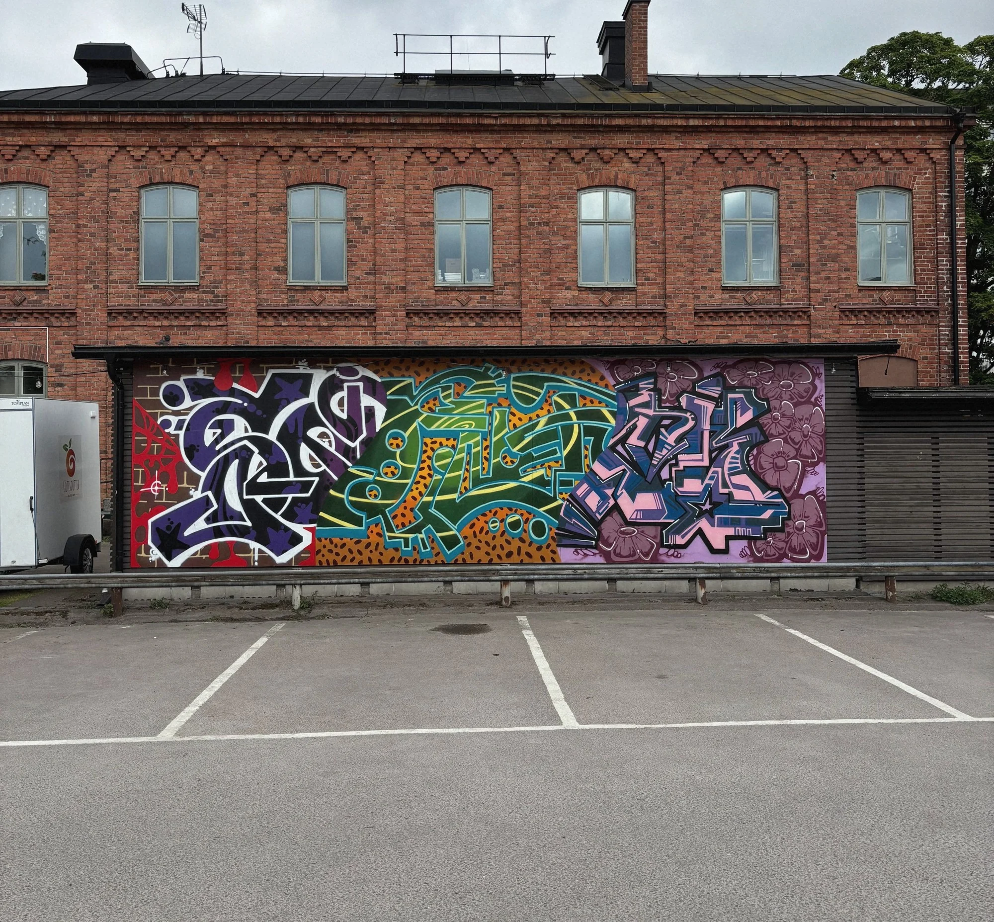 Den färdiga muralen Lager på lager i tre fält på en vägg framför en tegelbyggnad, med en parkering i förgrunden