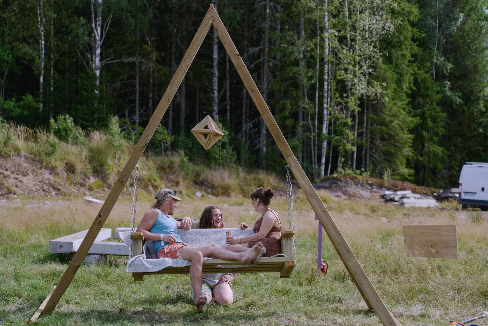 Besökare i en stor gunga under en triangulär träkonstruktion på en äng vid skogskanten