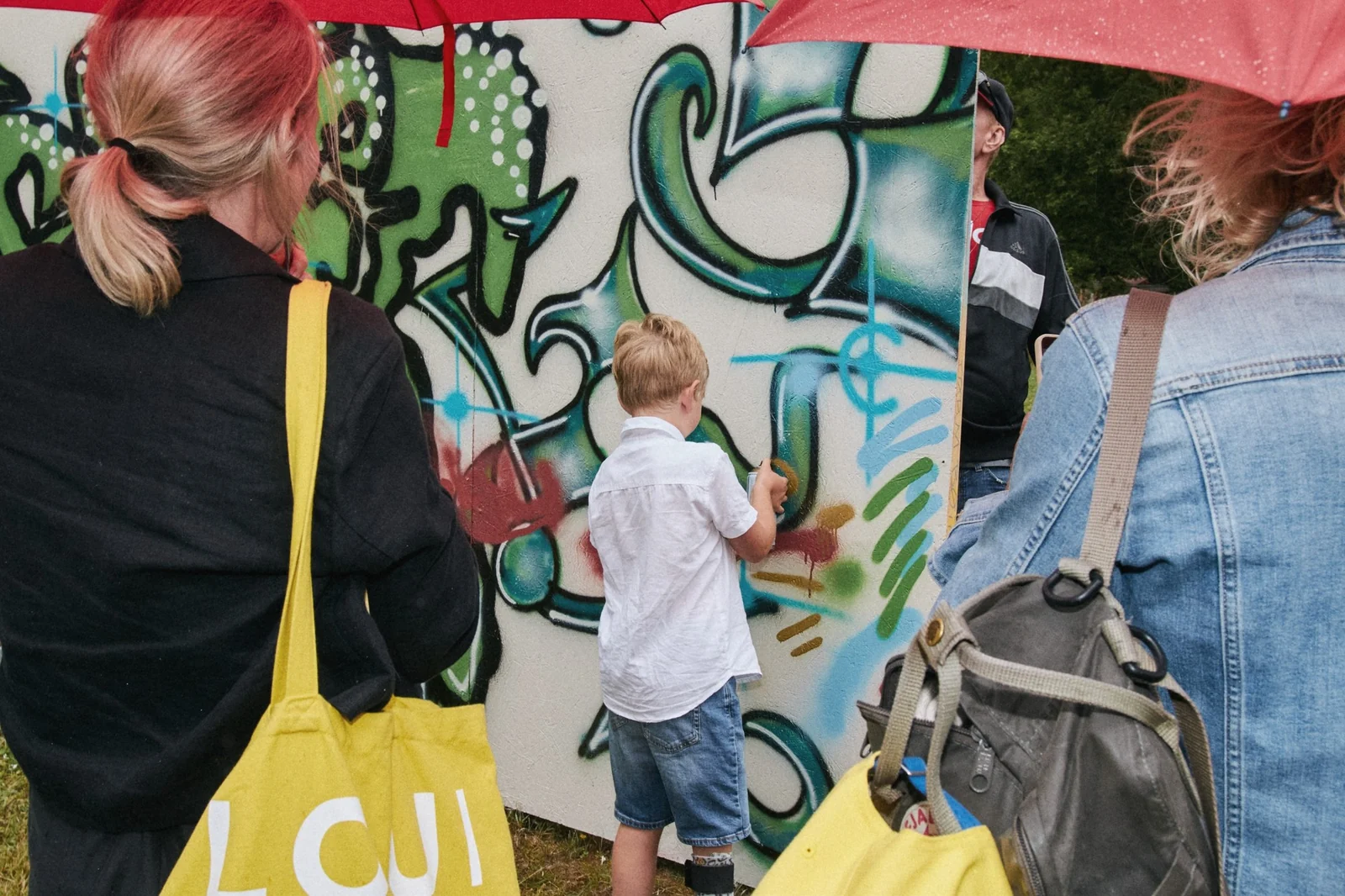 Ett barn sprayar på en graffitivägg medan vuxna med paraplyer står runt omkring, sedda bakifrån