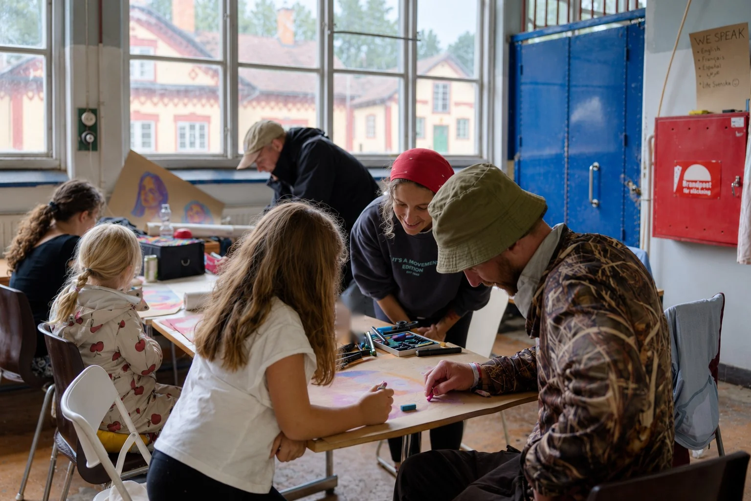Deltagare i olika åldrar skissar med kritor vid ett bord, barnen sedda bakifrån